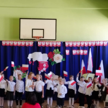 Grupa dzieci ubranych na galowo z flagami, prezentująca się na scenie podczas szkolnego wydarzenia.